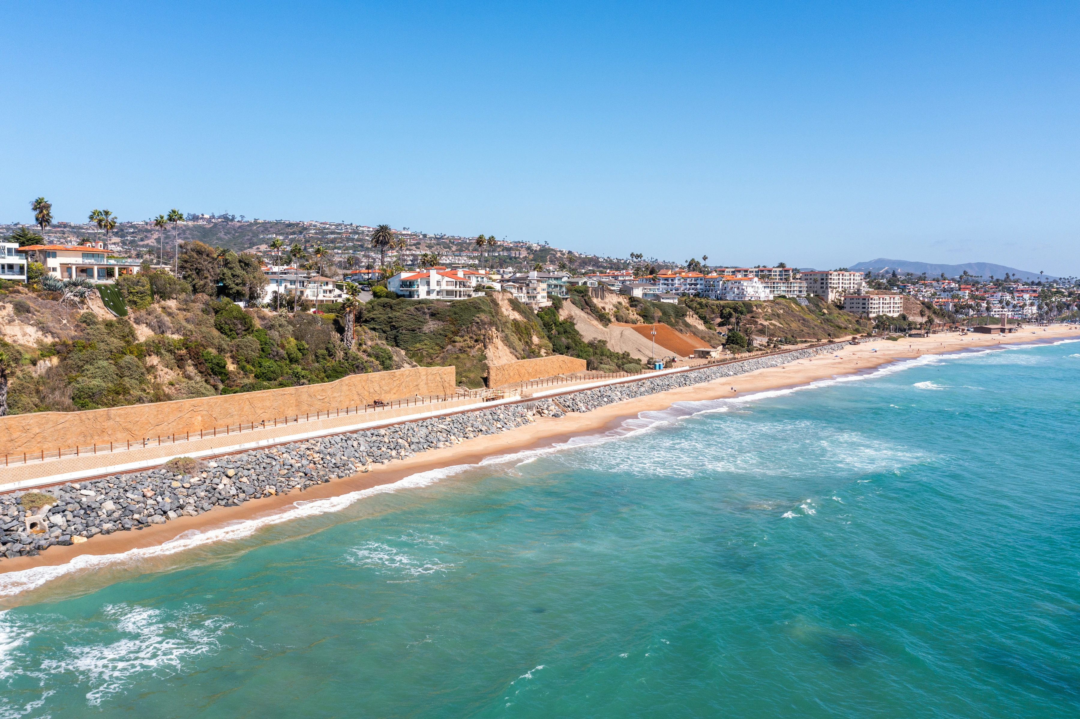Wide view of the coastal rail corridor with tan retaining-wall sections, graded bluff soil, and continuous gray shoreline rock.