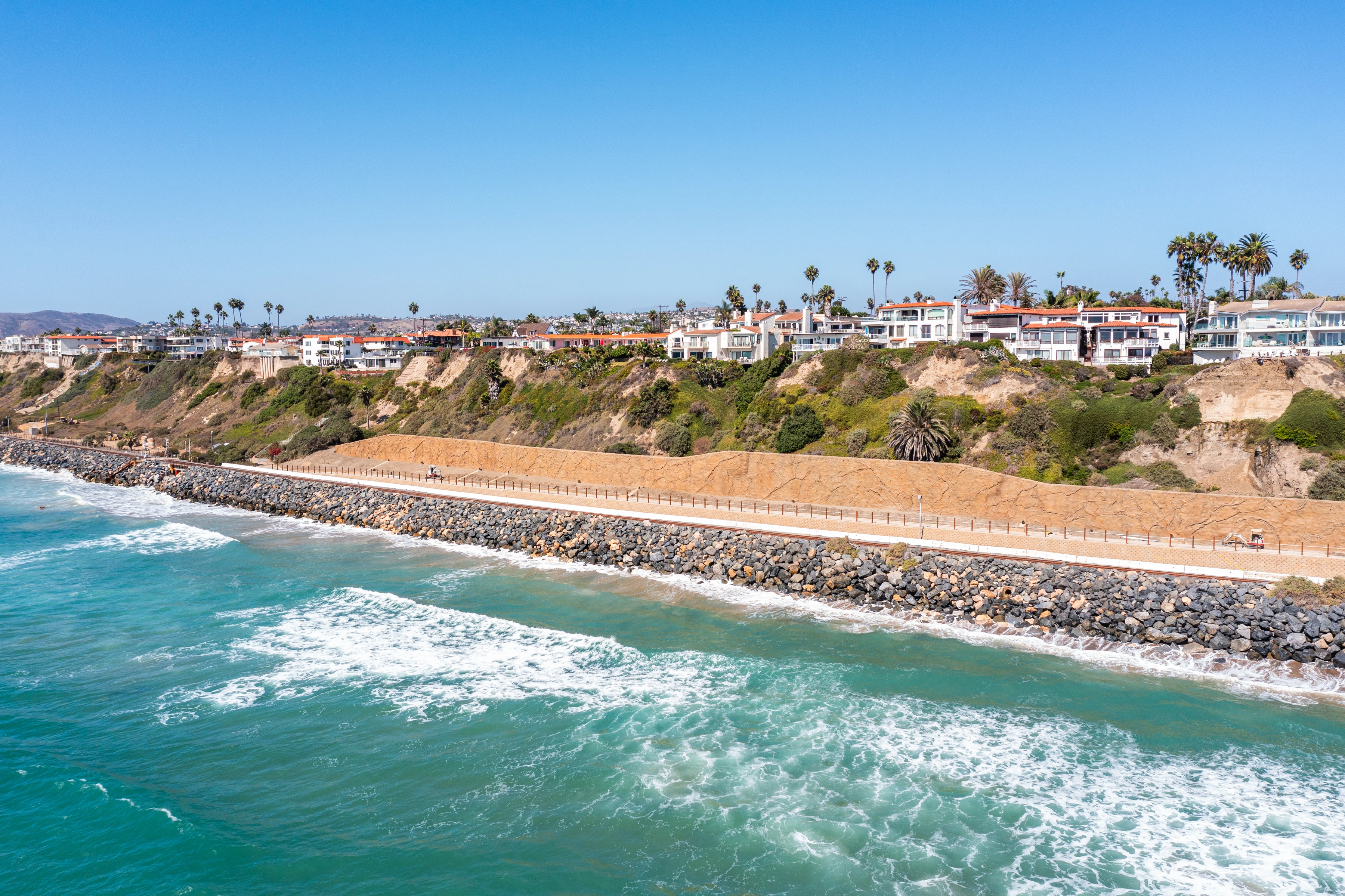 Long coastal view of shoreline riprap, rail lines, a textured retaining structure, and closely spaced posts below the bluff.