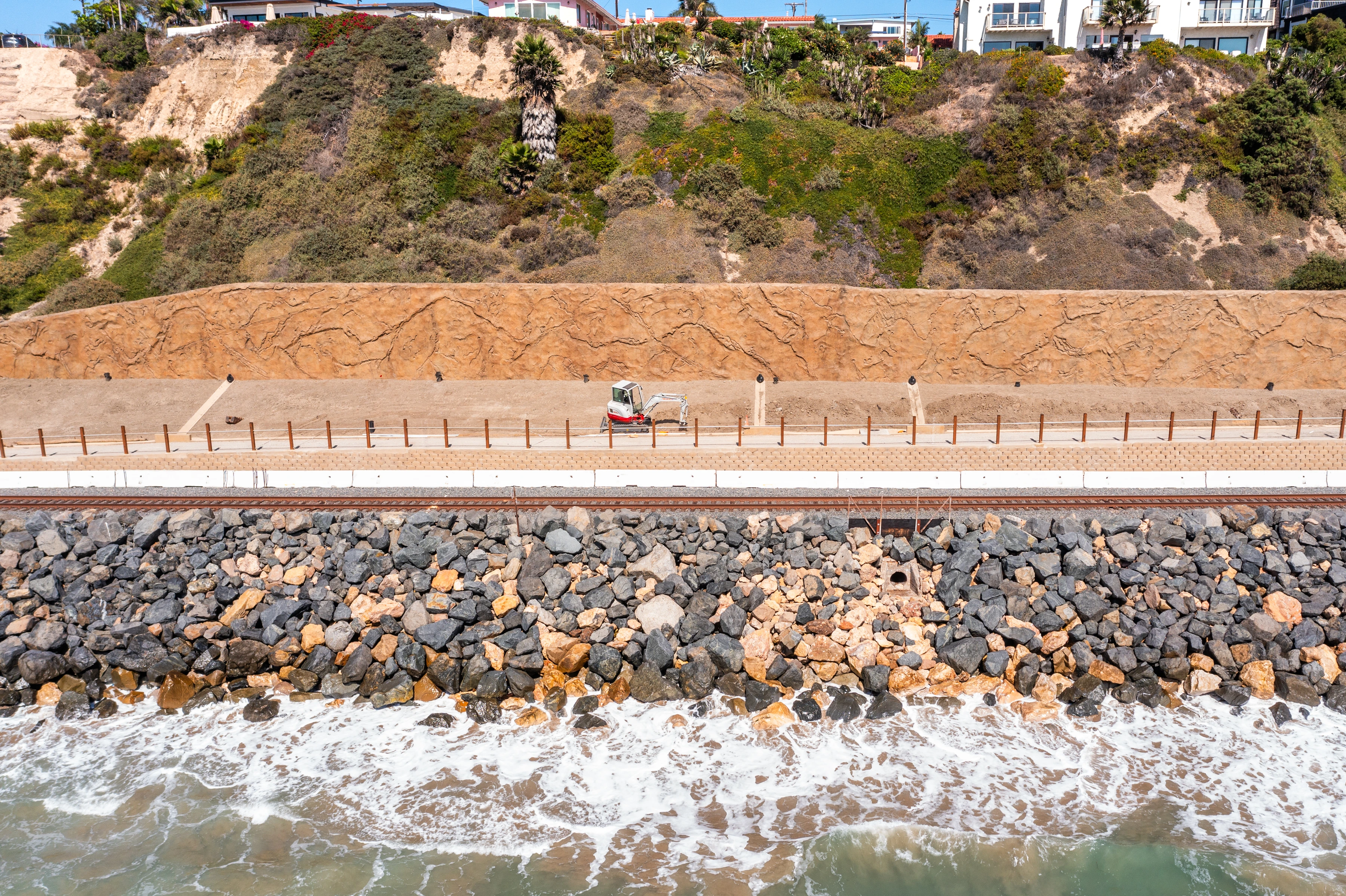 Compact tracked excavator on a graded strip beside a textured retaining wall, rail tracks, concrete elements, and shoreline armoring.