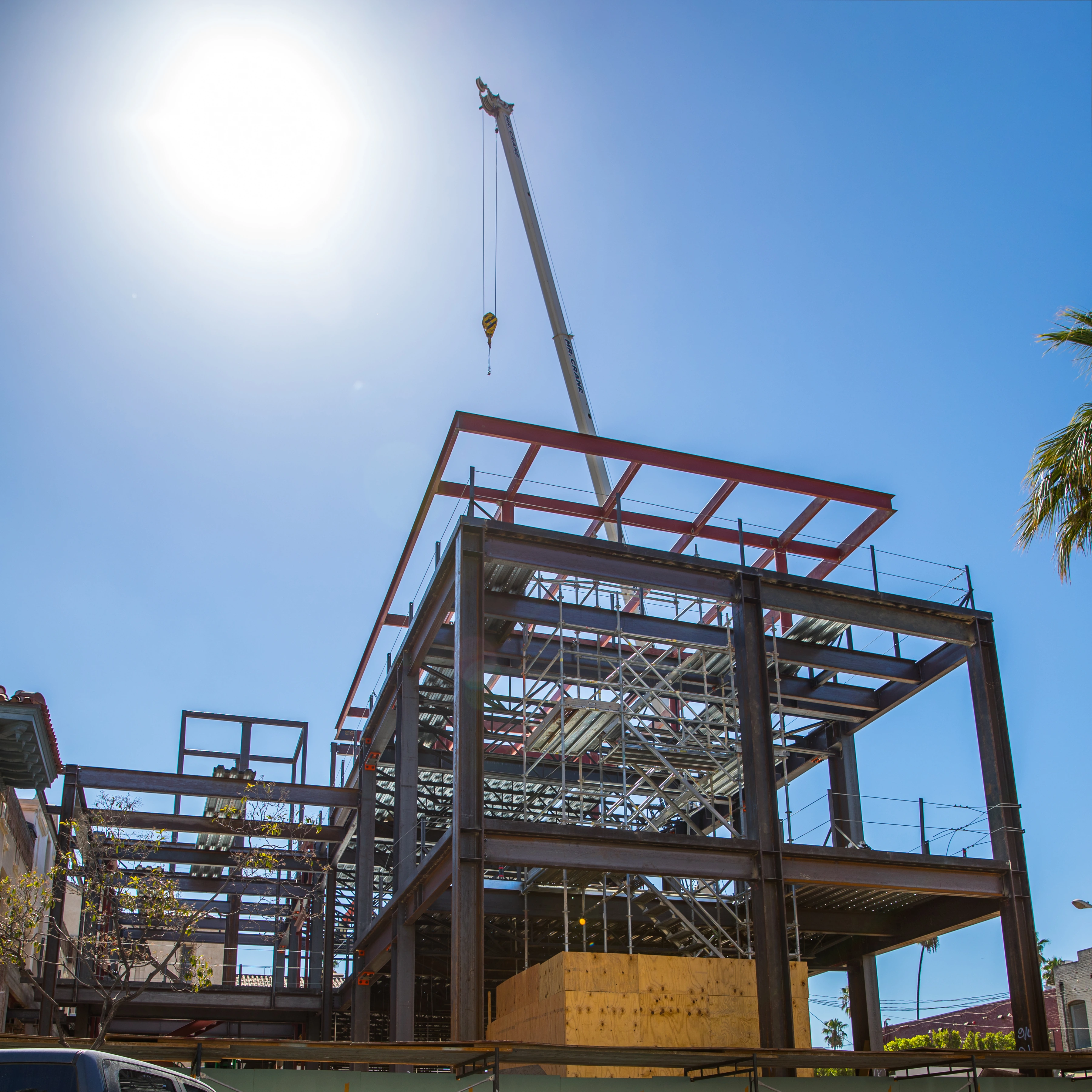 Low-angle view of a multistory steel frame with partial metal decking, interior scaffold, edge protection, and a crane hook overhead.