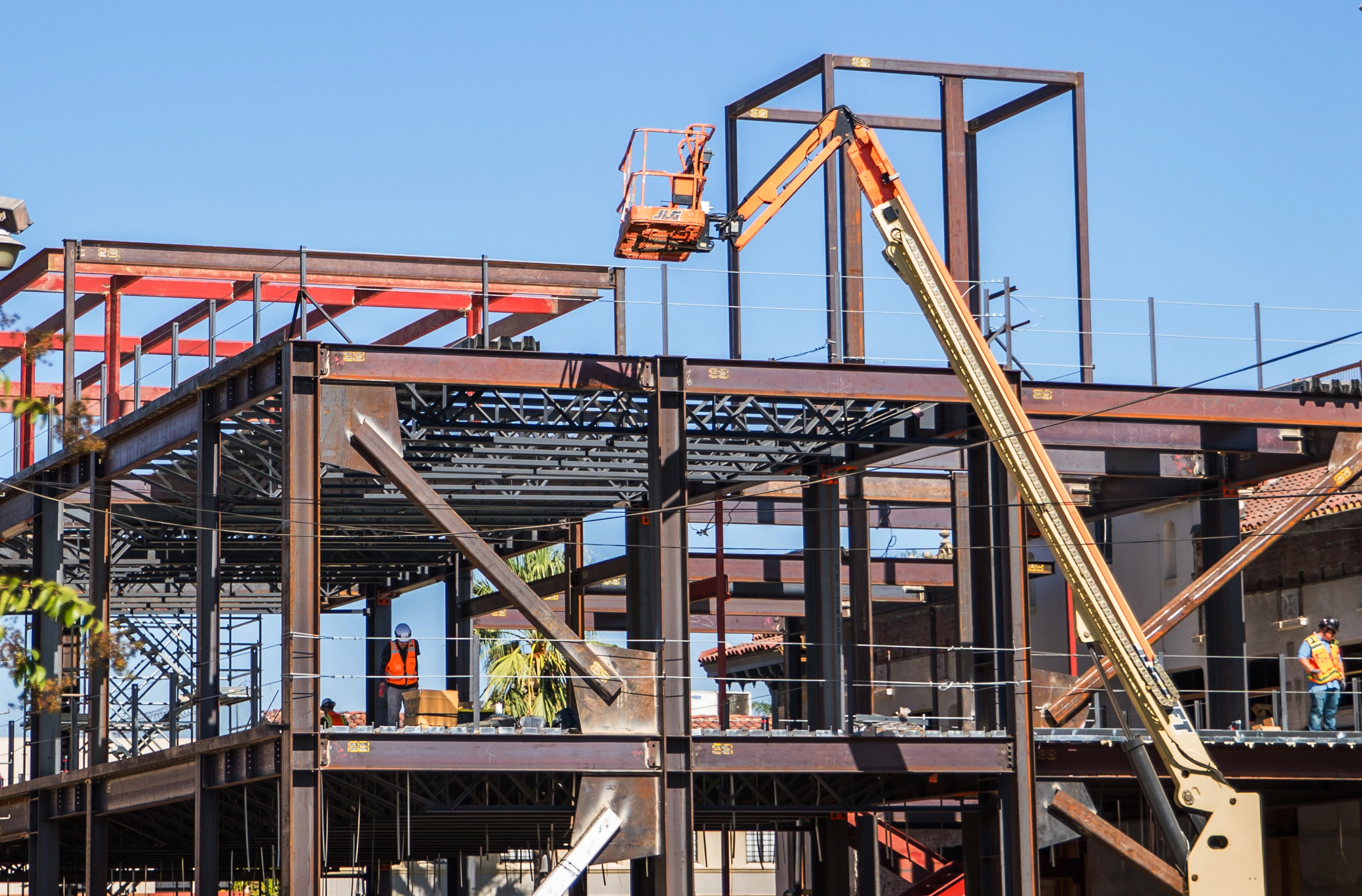 Exposed multilevel steel framing with an elevated boom lift, open web joists, safety cables, and workers in high-visibility gear.