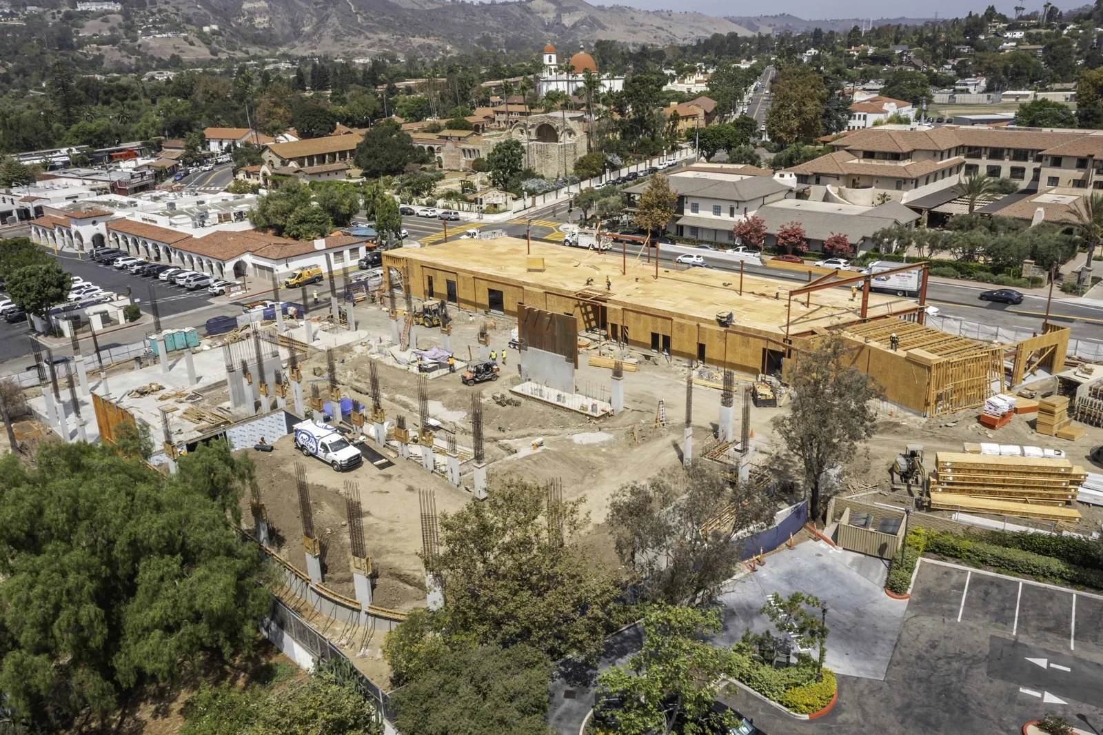 Wide construction view with concrete columns in the foreground, a wood roof deck beyond, and staged materials and equipment across the site.