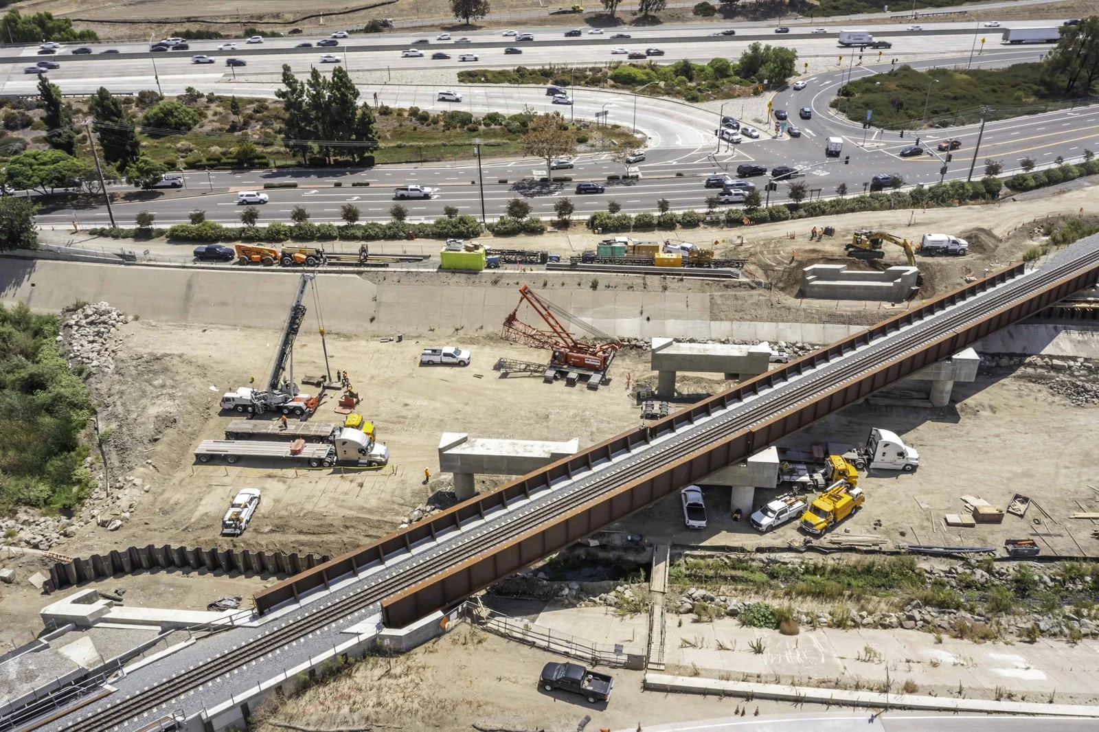 Elevated view of two rail tracks on the steel bridge with concrete supports, cranes, trucks and graded soil below.