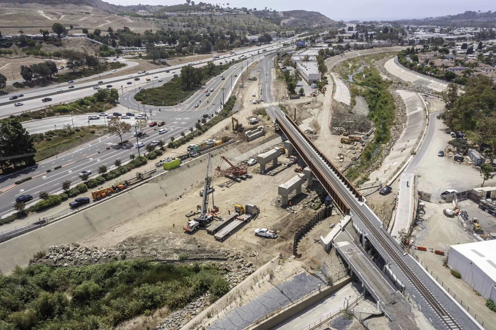 Wide view of the San Juan Creek rail crossing with steel girders, concrete columns, heavy equipment and nearby roads.
