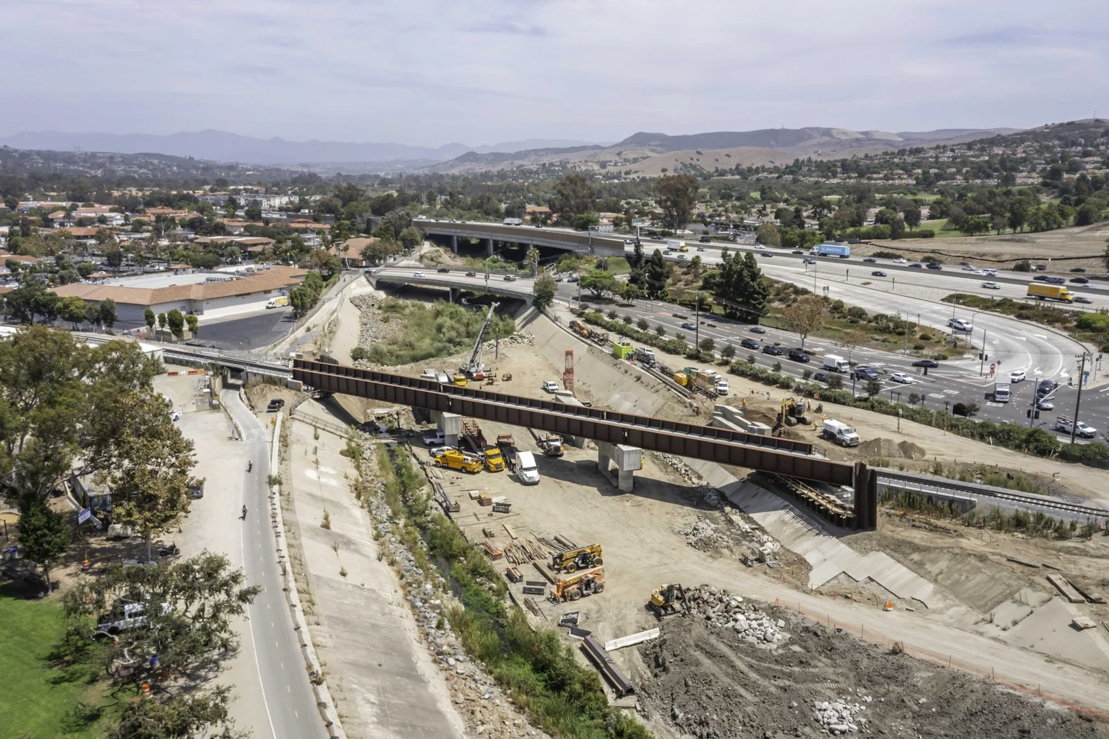 Wide elevated view of the long steel bridge over the mostly dry channel, with earthwork, equipment, rock and staged materials.