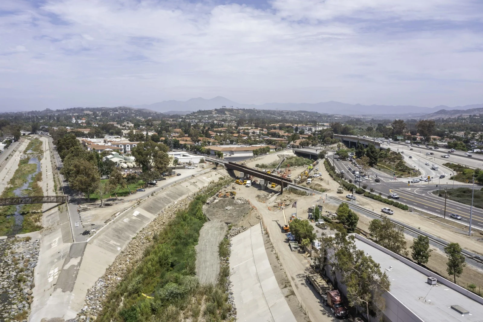 Elevated overview of the steel girder bridge, rail tracks, channel earthwork and nearby multilane interchange in San Juan Capistrano.